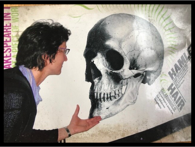 A person with short dark hair holding their hand up to an enlarged image of a skull, as if holding it 