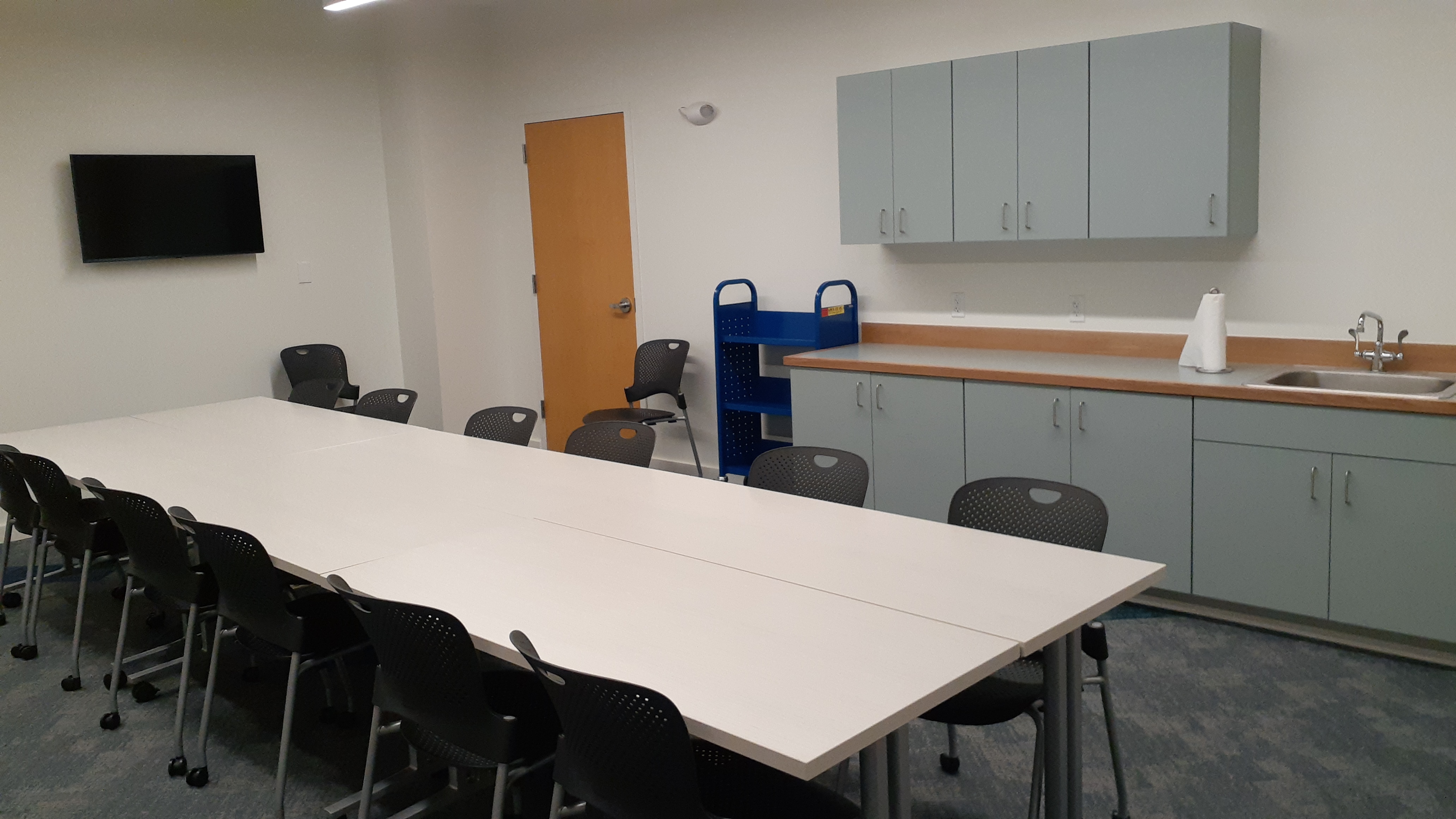 Image of the makerspace including a large conference-style table with twelve chairs, wall-mounted television, cabinets, and sink space